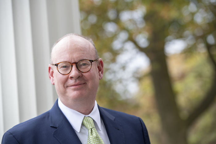 image of Penn State Schuylkill Chancellor Patrick M Jones standing outdoors during the fall with changing leaves in the distance and a white column nearby on the left of the frame